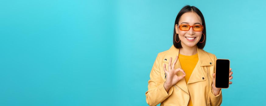 Smiling korean woman showing mobile phone app interface, smartphone application, recommending on cellphone, standing over blue background.