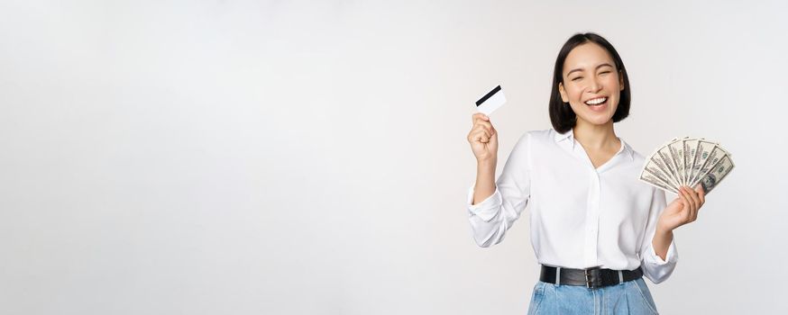 Happy korean woman holding credit card and money dollars, smiling and laughing, posing against white studio background.