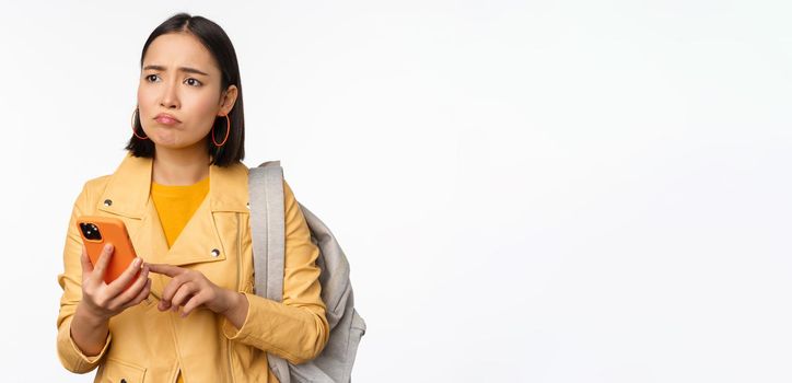 Image of happy girl traveller, tourist with backpack looking at smartphone, using mobile phone route app, standing over white background. Copy space