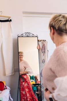 mature blonde woman, dressed in long red skirt and pink jumper. trying on clothes in front of the mirror in a clothing shop. shopping concept, person enjoying a shopping day. shopping concept. clothing saleswoman in her small shop. natural light from window, sunbeams, mirror, display with clothes, clothes rack, window displays, clothes, Vertical.