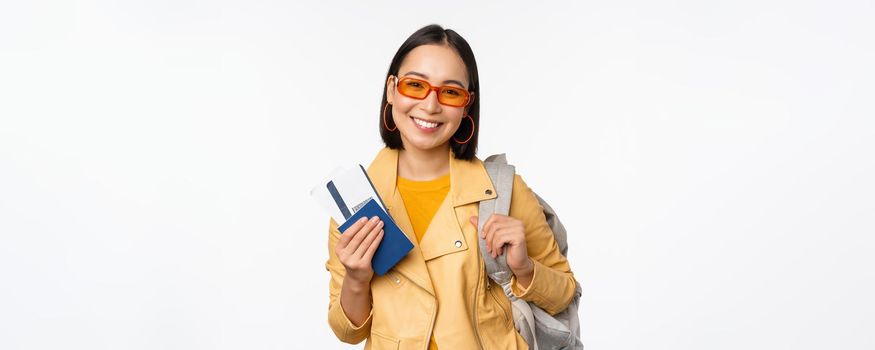 Happy asian girl going on vacation, holding passport and flight tickets, backpack on shoulder. Young woman tourist travelling abroad, standing over white background.
