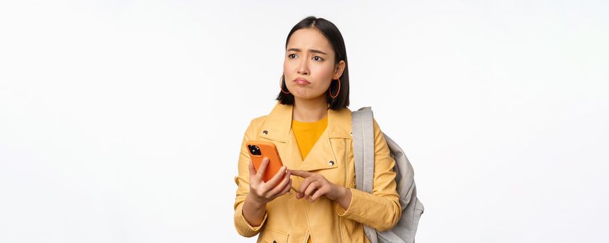 Image of happy girl traveller, tourist with backpack looking at smartphone, using mobile phone route app, standing over white background. Copy space