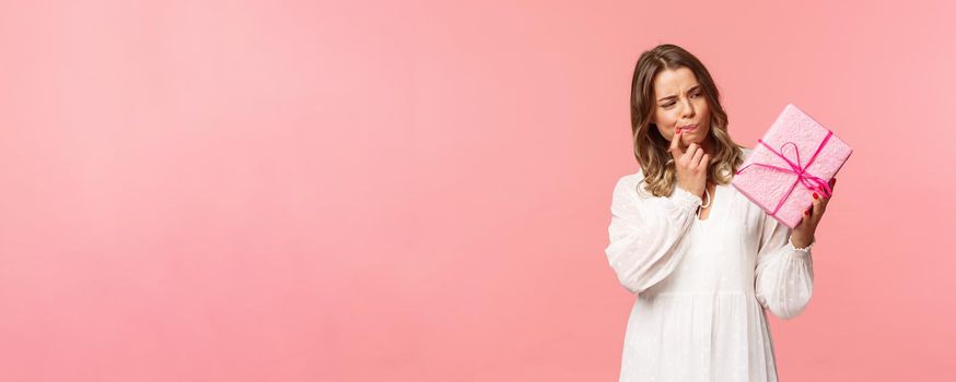 Holidays, celebration and women concept. Portrait of thoughtful curious blond girl thinking whats inside gift box, look pondering say hmm as making assumpion, standing pink background.