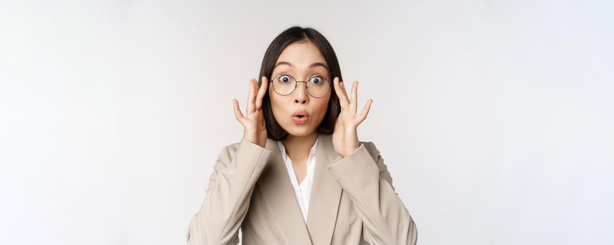 Portrait of asian businesswoman in glasses, looking surprised amazed at camera, standing in suit over white background.