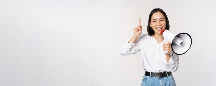 Smiling happy asian girl talking in megaphone and pointing up, announcing discount promo, showing advertisement on top, standing over white background.