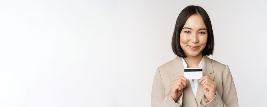 Smiling office clerk, asian corporate woman showing credit card, standing over white background in beige suit. Copy space