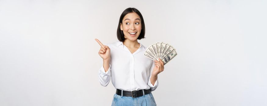 Smiling young modern asian woman, pointing at banner advertisement, holding cash money dollars, standing over white background.