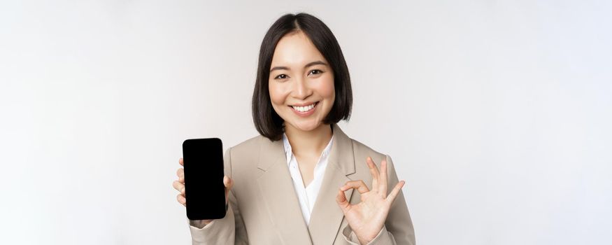 Smiling asian woman showing smartphone screen and okay sign. Corporate person demonstrates mobile phone app interface, standing over white background.