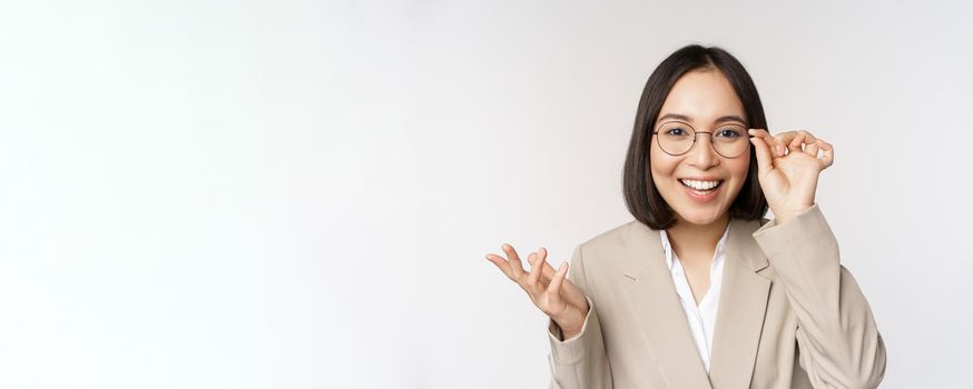 Enthusiastic asian saleswoman in glasses, smiling and laughing, looking amazed at camera, standing in beige suit over white background.