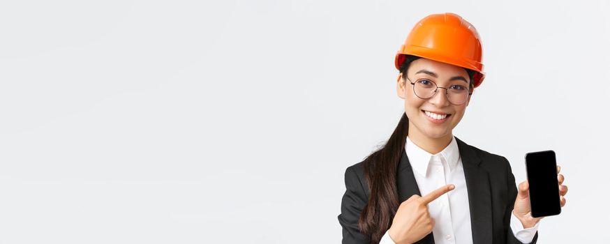 Close-up of smiling professional female asian construction engineer, architect in suit and safety mask introduce application, pointing finger at smartphone display, white background.
