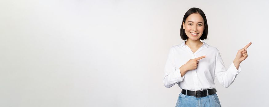 Portrait of beautiful asian woman, saleswoman pointing fingers right and showing info, banner or logo, sale advertisement, standing over white background.