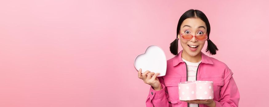 Cute asian girl giving you gift in heart shaped box, kissing and smiling, concept of holiday and celebration, standing over pink background.