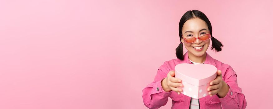 Cute asian girl giving you gift in heart shaped box, kissing and smiling, concept of holiday and celebration, standing over pink background.