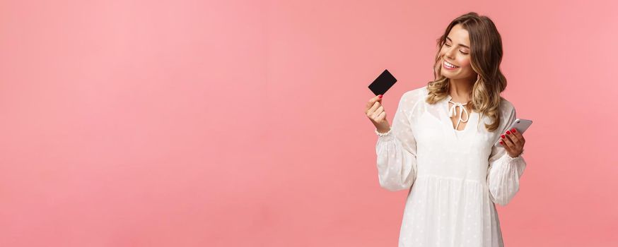 Portrait of happy good-looking young blond woman in white dress, looking pleased and satisfied at credit card, holding smartphone, recommend buy online, internet shopping concept.