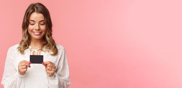 Close-up portrait of excited and amused blond girl in white dress, holding credit card and smiling thrilled, cant resist temptation to buy something, waste money online shopping, pink background.