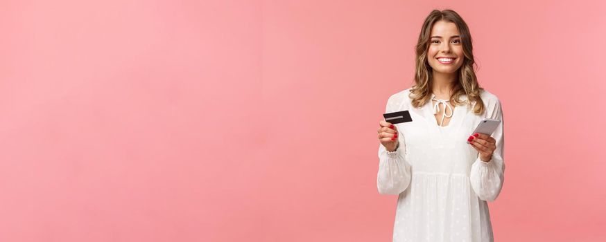 Portrait of happy smiling blond woman order food online, use application for shopping internet store, holding credit card and smartphone, grinning delighted at camera, pink background.