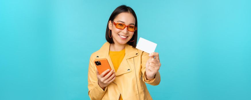 Online shopping. Stylish young asian woman in sunglasses, showing credit card and using smartphone, paying in internet, making purchase, standing over blue background.