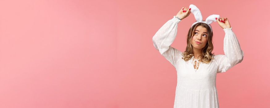 Holidays, spring and party concept. Portrait of silly cute blond girl in rabbit ears, looking up daydreaming, smiling lovely wear white dress, standing over pink background carefree.