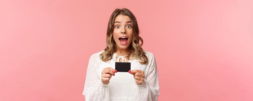 Close-up portrait of emotive, excited and thrilled good-looking blond girl showing credit card and feeling very happy, telling about discounts, special offers, bank service, pink background.