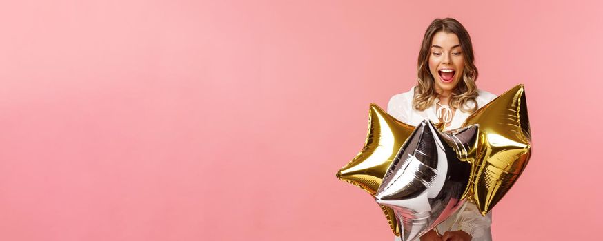 Holidays, celebration and women concept. Portrait of joyful beautiful blond girl in white dress being excited and impressed with cool start-shaped balloons, partying over pink background.