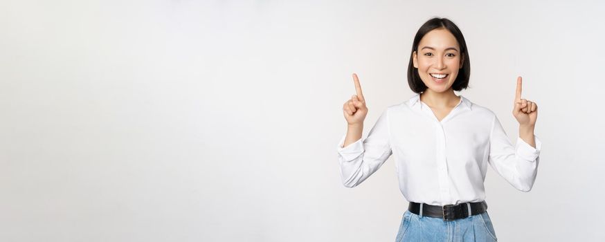 Image of smiling confident businesswoman, asian lady pointing fingers up, showing banner or sale info, standing over white background.