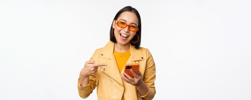 Happy smiling korean girl in sunglasses, holding smartphone, pointing at mobile phone screen, standing over white background. Copy space