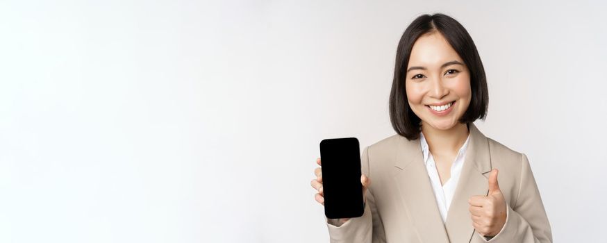 Smiling asian woman showing smartphone screen and thumbs up. Corporate person demonstrates mobile phone app interface, standing over white background.
