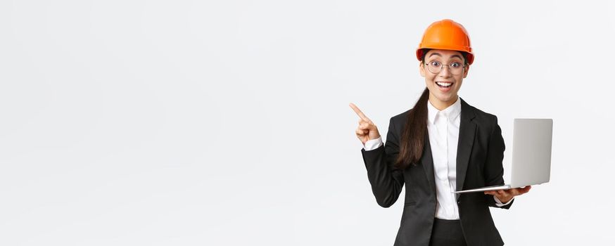 Excited happy asian female engineer, industrial woman in safety helmet and business suit, showing presentation, pointing finger at graph or chart and holding laptop computer, smiling amazed.