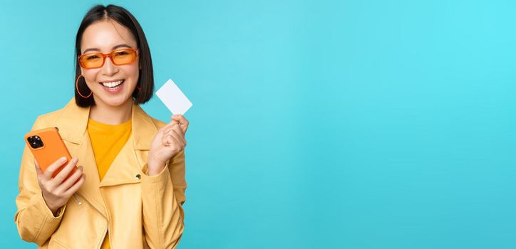 Online shopping. Stylish young asian woman in sunglasses, showing credit card and using smartphone, paying in internet, making purchase, standing over blue background.