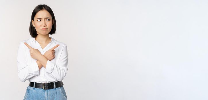 Indecisive asian woman pointing fingers sideways, pointing fingers and looking clueless, confused with choices, standing over white background.