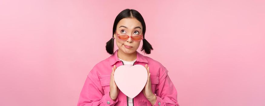 Beautiful asian girl smiling happy, showing heart gift box and looking excited at camera, standing over pink romantic background.