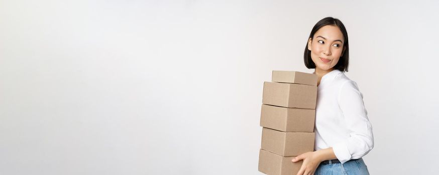 Portrait of young asian woman holding boxes, carry delivery goods. Korean female entrepreneur assemble order, standing voer white background.