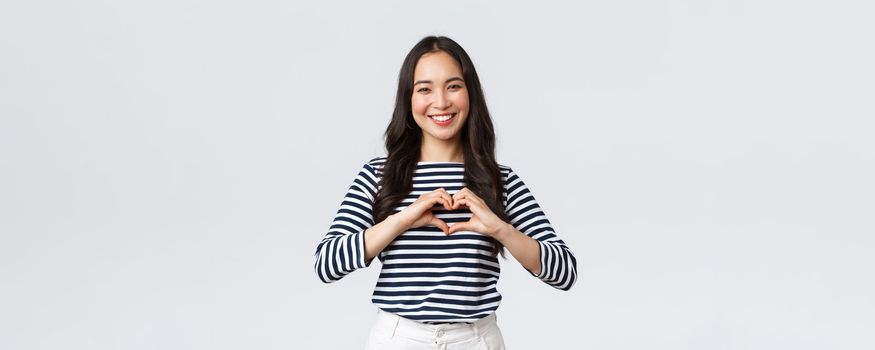 Lifestyle, people emotions and casual concept. Lovely smiling adorable asian woman showing heart sign and smiling, express sympathy or care, standing white background.