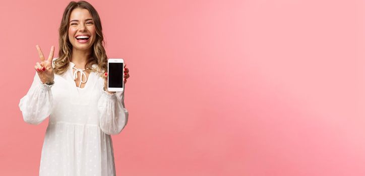 Portrait of kawaii optimistic and happy young girl in white dress, show mobile phone display and peace sign, laughing feeling cheerful and glad to share awesome app, useful link, pink background.