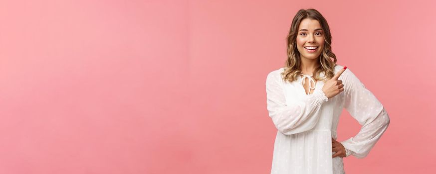 Upbeat good-looking caucasian blond girl in white dress, pointing upper right corner and smiling at camera with interested, curious face discuss special promo spring discounts, pink background.