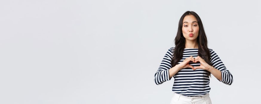 Lifestyle, people emotions and casual concept. Lovely smiling adorable asian woman showing heart sign and smiling, express sympathy or care, sending air kiss at camera, standing white background.