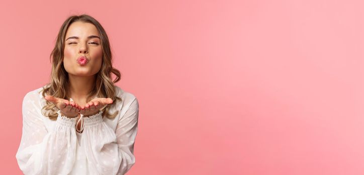 Close-up portrait of lovely feminine girlfriend enjoying spring fine day, sending air kiss at camera, winking and folding lips in mwah, hold hands near mouth, pink background.