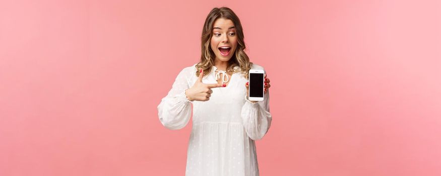 Portrait of excited and amazed, happy blond female in white dress, pointing finger at mobile phone screen, looking at smartphone with pleased and surprised smile, stand pink background.