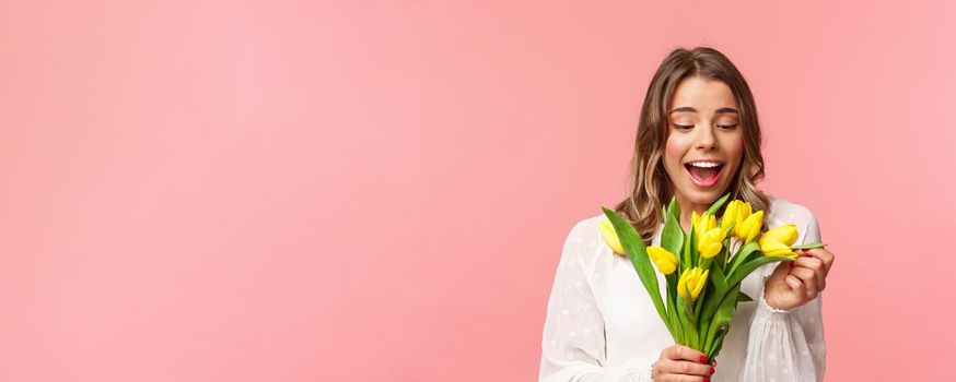 Spring, happiness and celebration concept. Portrait of surprised and pleased happy girl cheerfully looking at bouquet of flowers, smiling amused, receive yellow tulips, stand pink background.