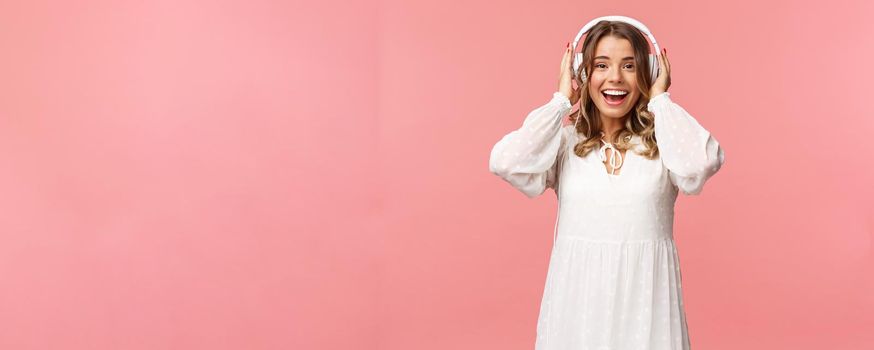 Portrait of excited, happy good-looking girl in white tender dress, wearing headphones and smiling amazed as looking at camera, fascinated with good sound quality, pink background.