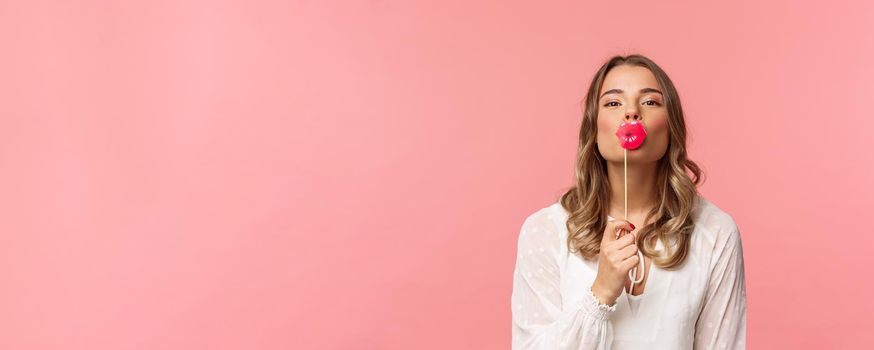 Spring, happiness and celebration concept. Close-up of romantic attractive blond caucasian girl in white dress, holding big kissing lips stick over mouth and looking lovely camera, pink background.