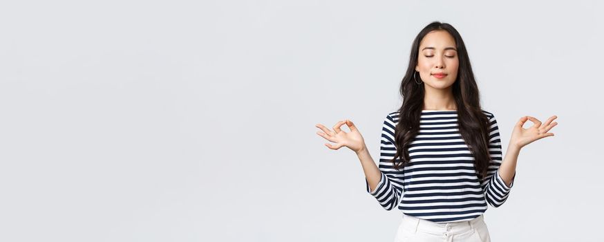 Lifestyle, people emotions and casual concept. Relaxed and patient smiling young asian woman with closed eyes meditating to calm down, do breathing exercises with hands in zen gesture.