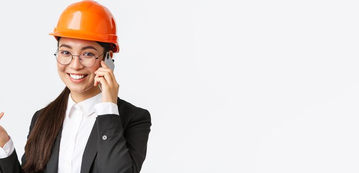 Close-up of asian businesswoman manage enterprise, engineer in safety helmet and suit having phone conversation, calling investors, smiling while talking over smartphone, white background.