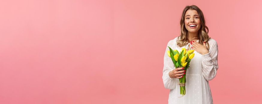 Holidays, beauty and spring concept. Grateful charming blond girl receive yellow tulips, feel touched thanking for romantic gift, sighing lovely smiling, feel happy hold flowers, pink background.