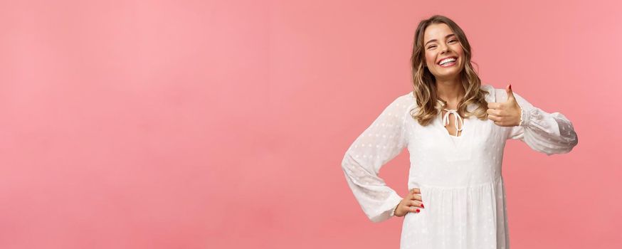 Enthusiastic lovely young woman in white cute dress, show thumb-up in approval, recommend product, smiling satisfied, leave positive review, impression, standing pink background.