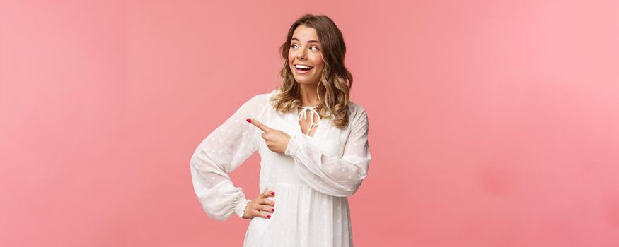 Curious upbeat beautiful blond woman enjoying summer feeling cheerful, pointing looking left with interested, enthusiastic expression, wear cute white dress, standing pink background.