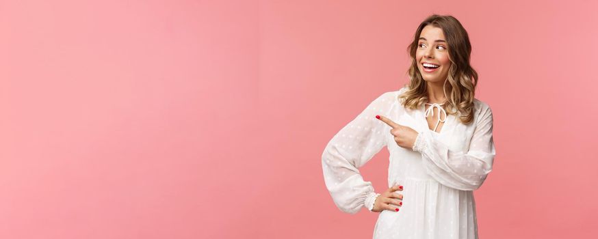 Curious upbeat beautiful blond woman enjoying summer feeling cheerful, pointing looking left with interested, enthusiastic expression, wear cute white dress, standing pink background.