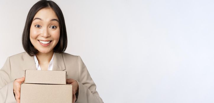 Image of asian saleswoman, business woman giving boxes with order, deliver to customer, standing in suit over white background.