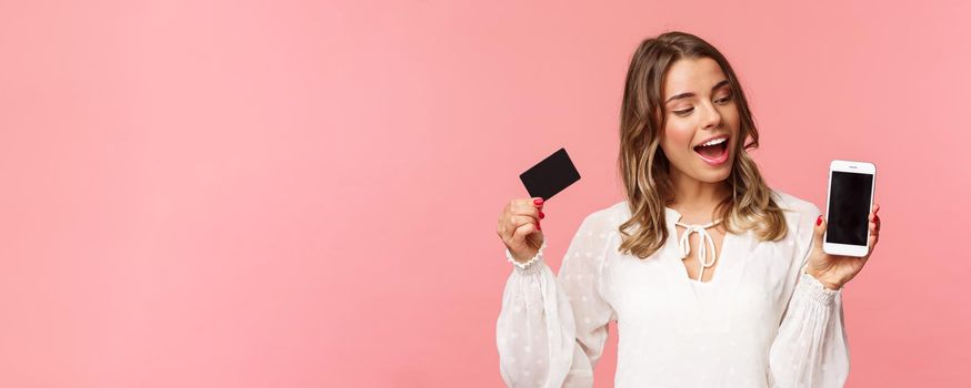 Finance, shopping and technology concept. Close-up portrait of excited good-looking blond girl in white dress, renew her spring wardrobe with buying clothes online via smartphone, hold credit card.