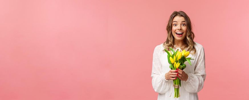 Holidays, beauty and spring concept. Portrait of excited and amazed young beautiful blond girl in white dress, holding yellow tulips, receive flowers as gift, smling amused, pink background.